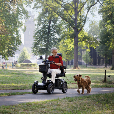 Ein Mann fährt auf seinem E-Mobil durch den Park mit seinem Hund an der Leine.