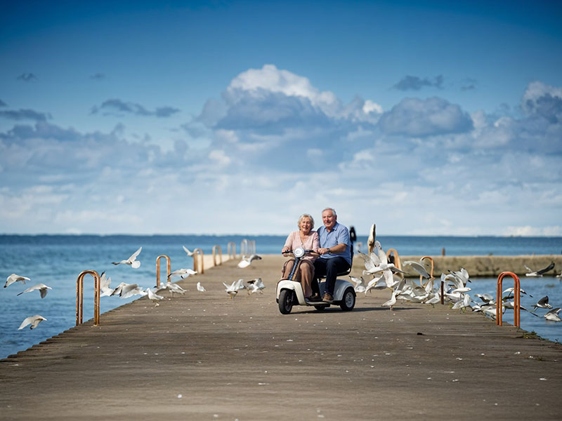 Ein älteres Pärchen sitzt zusammen auf einem Elektromobil am Holzsteg auf einer Strandpromenade. width=